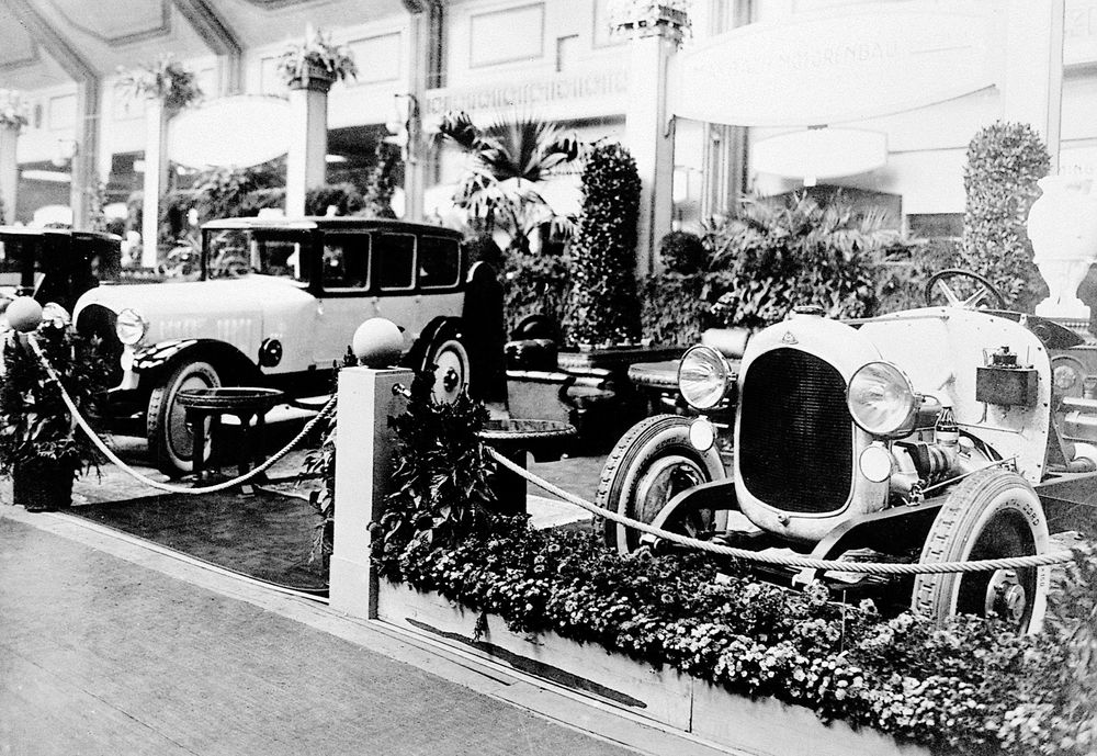 Die historische Aufnahme zeigt zwei Maybach Automobile auf einem Messegelände. Um die beiden Autos herum stehen einige Pflanzen und Blumen. 