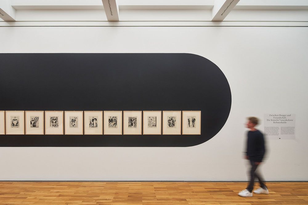 Die Ausstellungsansicht zeigt eine Reihe von Bildern die gleichmäßig nebeneinander an der Wand hängen. Daneben sieht man die Silhouette eines Mannes in Bewegung.