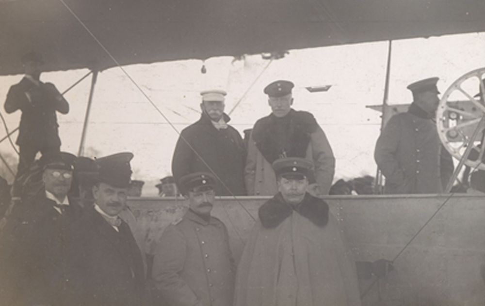 Die historische Aufnahme zeigt eine Gruppe von Männern, darunter Graf Zeppelin und Theodor Kober, die in und um eine Luftschiffgondel stehen.