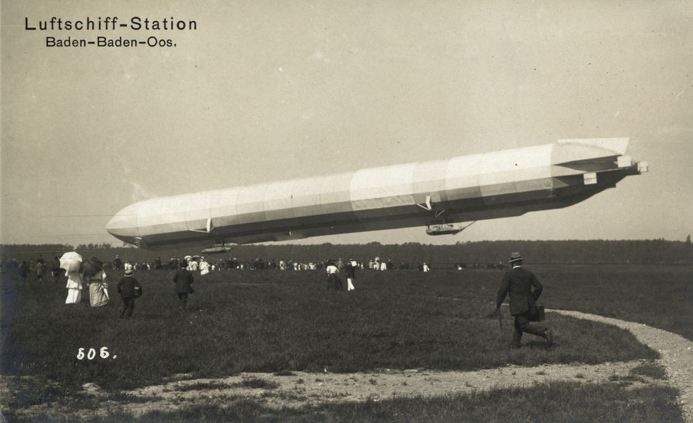 Die historische Aufnahme zeigt einen Zeppelin, der sich mit der Spitze kurz über dem Boden befindet. Mehrere Personen eilen in Richtung des Zeppelins.