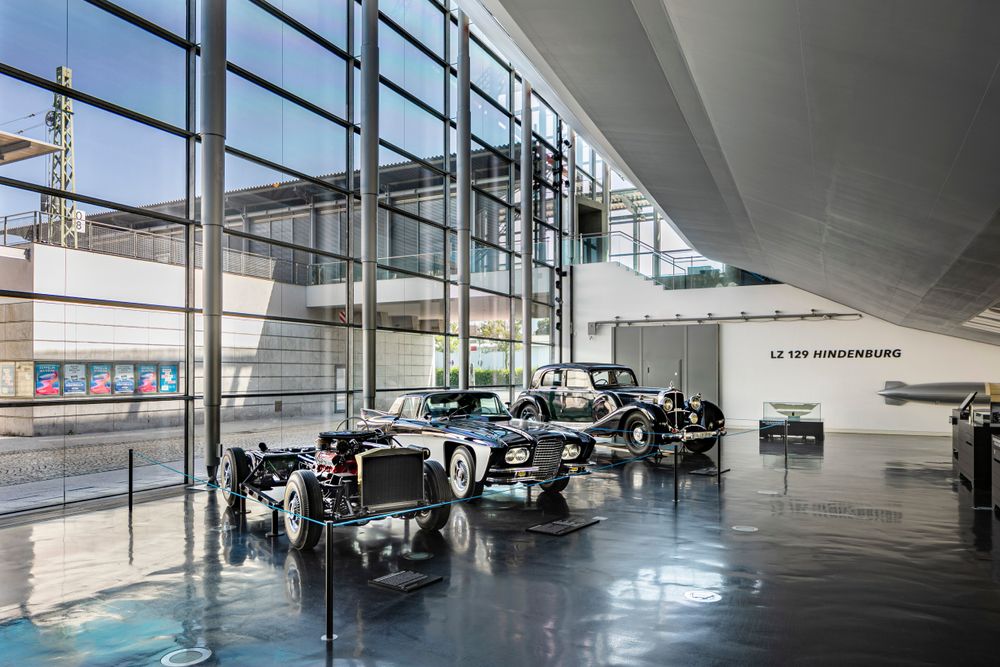 Ausstellungsansicht der Technikausstellung im Zeppelin Museum, drei historische Autos vor einer Glasfassade unter der Rekonstruktion des LZ 129 Hindenburg.