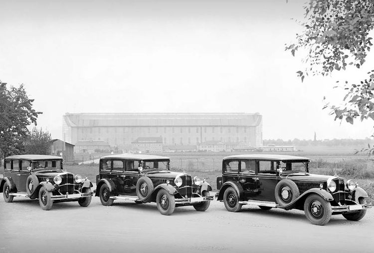 Die historische Aufnahme zeigt drei Maybach Automobile, die nebeneinander in einer Reihe stehen. Im Hintergrund sieht man eine riesige Luftschiffhalle.