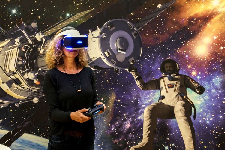 Eine Frau mit lockigen Haaren trägt eine VR-Brille und hält einen Gaming-Controller in den Händen. Hinter ihr an der Wand sind der Weltall, ein Raumschiff und ein Astronaut abgebildet.