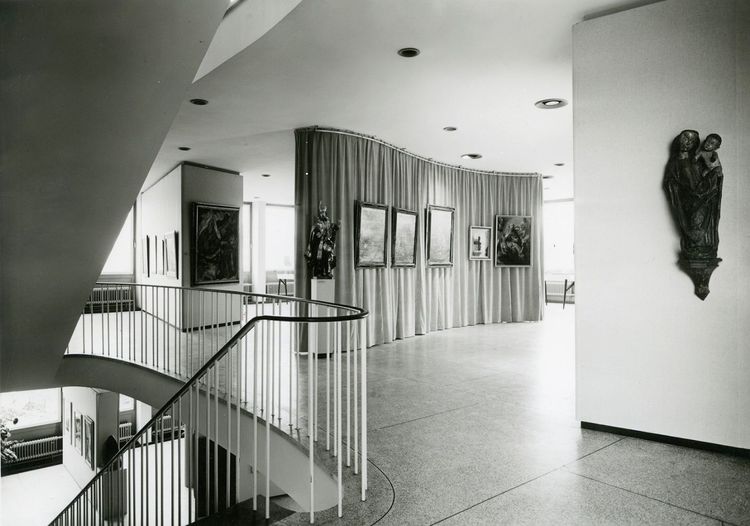 Die historische Aufnahme zeigt einen unterteilten Raum, in dem Gemälde an den Wänden hängen. Links befindet sich eine Treppe die weiter in das darüberliegende Geschoss führt. 