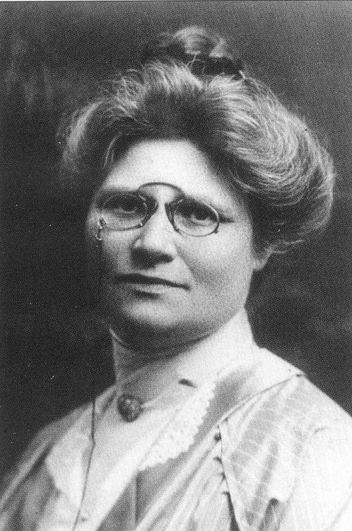 Die historische Aufnahme zeigt das Porträt einer Frau mit auffälliger Hochsteckfrisur. Die Frau trägt eine kleine Brille, an der eine Schnur hängt.