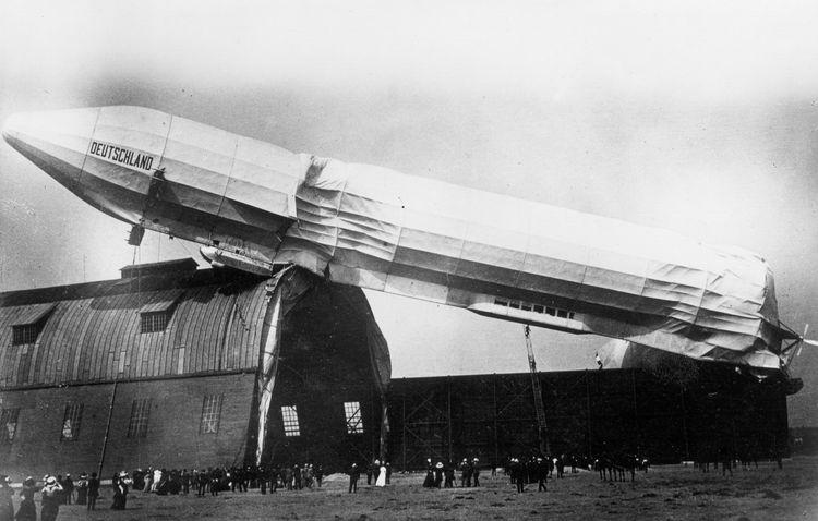 Die historische Aufnahme zeigt ein Luftschiff, das gegen eine Luftschiffhalle prallt und beschädigt wird. Eine große Menge an Personen steht außerhalb der Halle auf dem Feld und beobachtet das Geschehen.