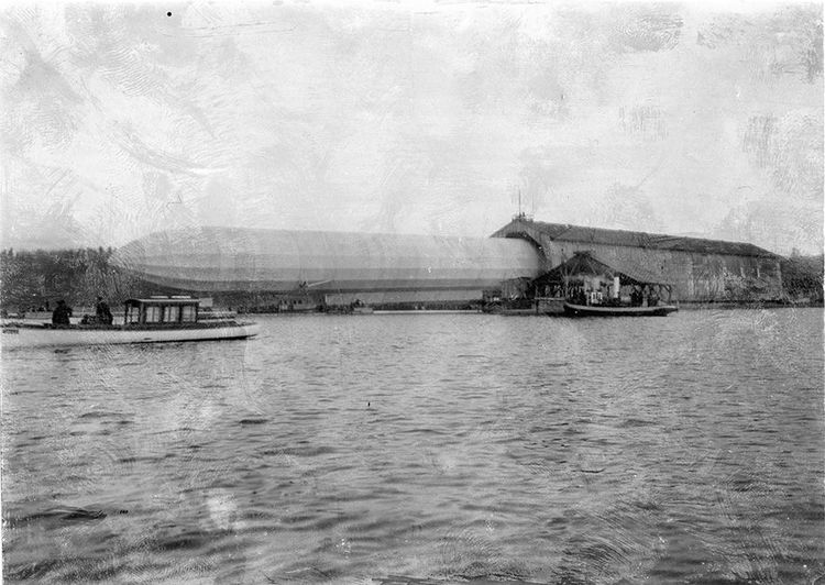 LZ 2 beim Aushallen aus der schwimmenden Halle in Manzell, im Mittelgrund der schwarz-weiß Fotografie. Dreiviertel des Luftschiffes sind schon zu sehen, links in der Bildmitte. Davor und vor der Halle sind zwei Motorboote sichtbar.