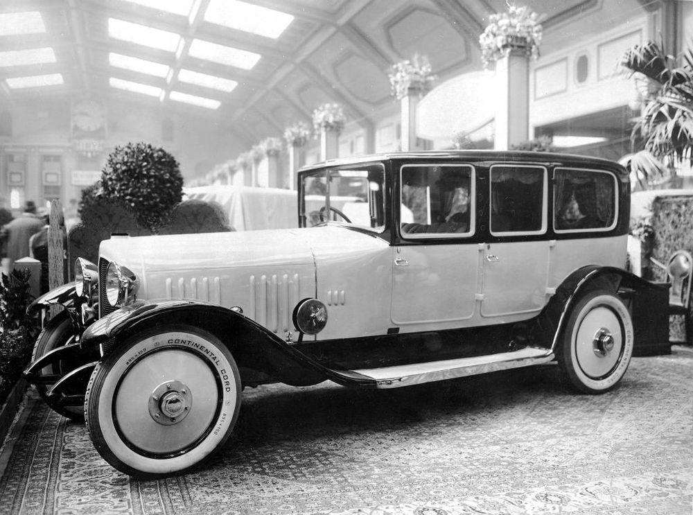 Die historische Aufnahme zeigt eine Herrenfahrerlimousine, die in einer Halle ausgestellt ist. Hinter dem Auto stehen Pflanzen und geschmückte Säulen. 