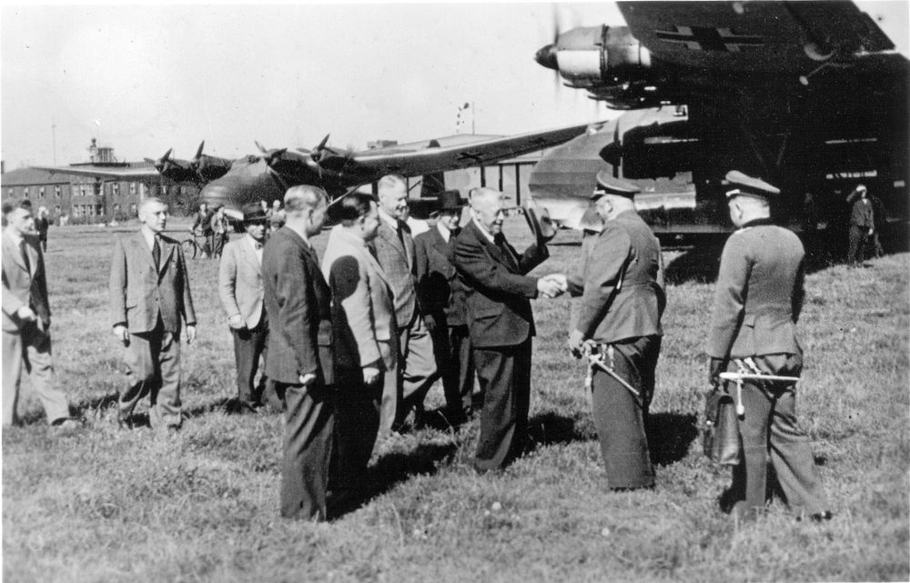 Eine Gruppe von Männern begegnet zwei Uniformierten. Hugo Eckener schüttelt einem der Uniformierten die Hand. Im Hintergrund sind Messerschmitt Gigant Flugzeuge sowie ein Flughafengelände zu sehen.