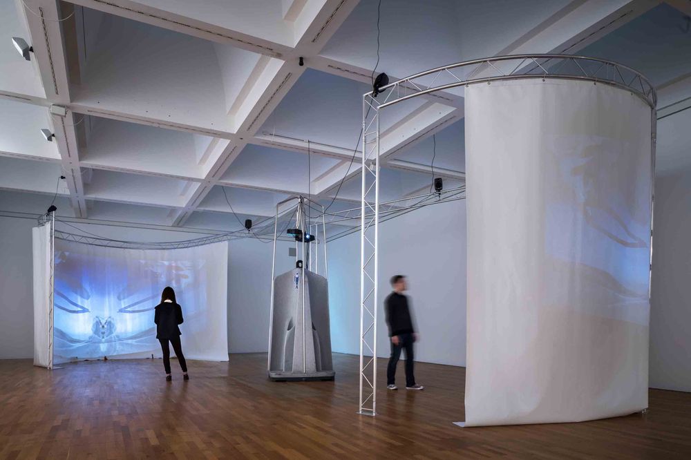 Die Ausstellungsansicht zeigt einen weißen Raum in dem im Halbkreis zwei sich gegenüberliegende Vorhänge aufgestellt sind. Beamer projizieren Videos auf die Vorhänge. 