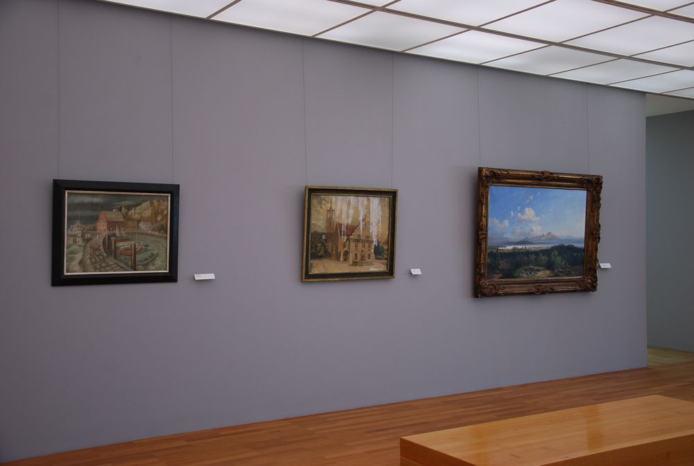 An einer grauen Wand hängen drei unterschiedliche Gemälde nebeneinander. Bei dem Bild in der Mitte verläuft weiße Farbe oder Flüssigkeit über dem Bild.