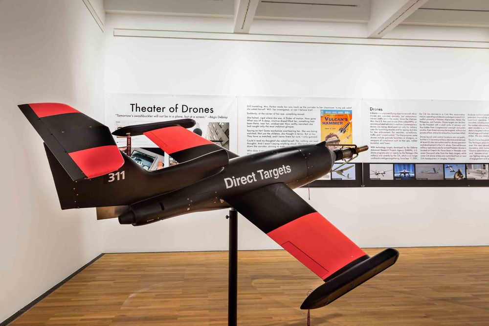 Die Ausstellungsansicht zeigt das Modell eines schwarz-roten Flugzeugs. Dahinter an einer weißen Wand hängen Ausstellungstexte und verschiedene Bilder.