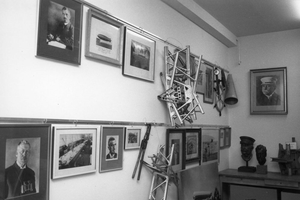 Schwarz-weiß Fotografie des ehemaligen Archivs. An den Wänden hängen Bilderrahmen mit Portraits und Fragmente von Dreiecksträgern.
