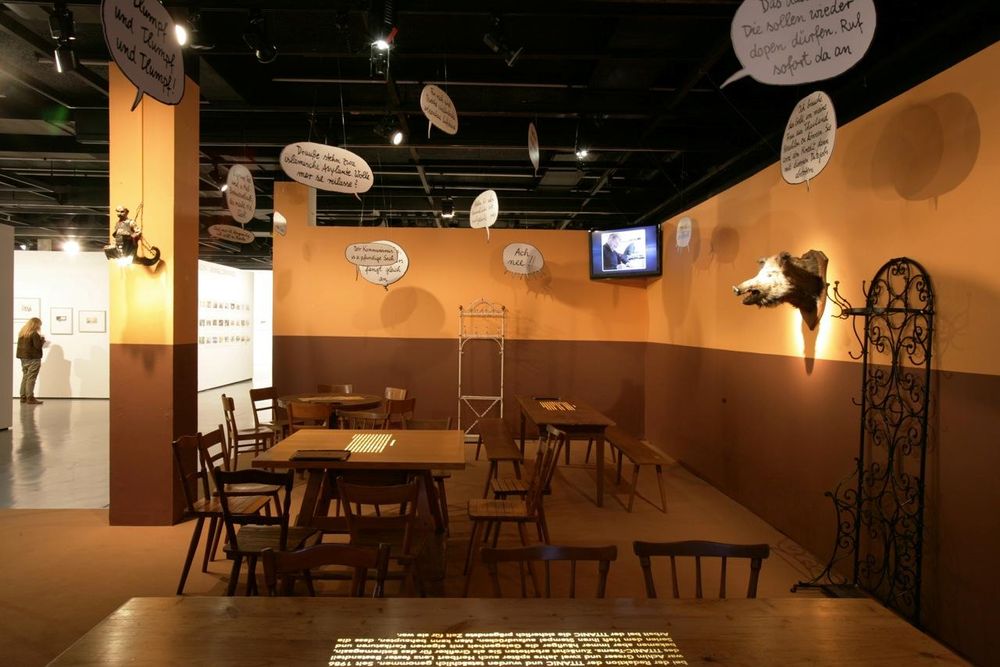 Der Ausstellungsraum ähnelt der Inneneinrichtung eines alten Gasthofes. Um massive Holztische stehen passende Holzstühle, rechts an der Wand hängt ein ausgestopfter Wildscheinkopf und von der Decke baumeln verschiedene Sprechblasen mit Text.