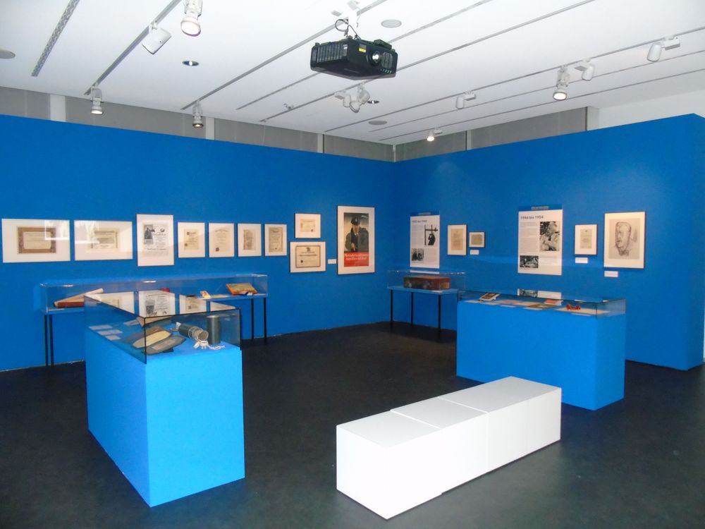 Die Ausstellungsansicht zeigt einen Raum mit blauen Wänden an dem verschiedene Bilder hängen. Zwischen zwei blauen Podesten mit Ausstellungsobjekten darauf steht eine weiße Sitzbank für Besucher*innen.