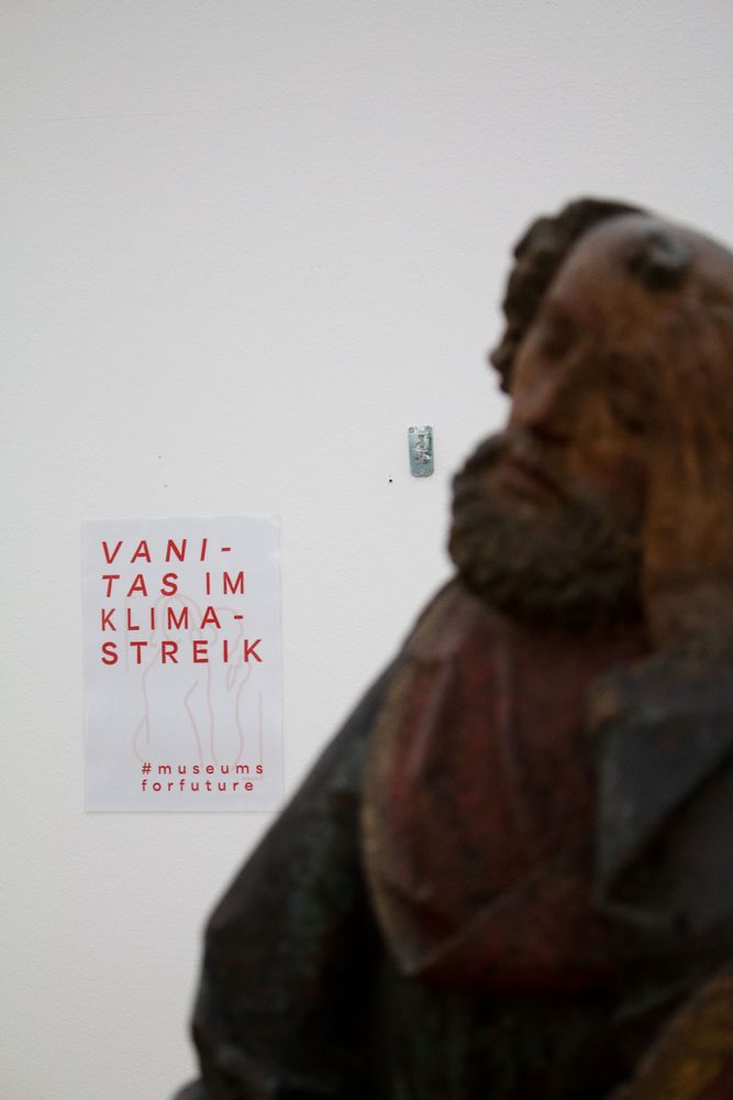 Im Hintergrund hängt an einer weißen Wand ein Plakat mit der Aufschrift "Vanitas im Klimastreick". Im Vordergrund ist eine Holzskulptur zu sehen.
