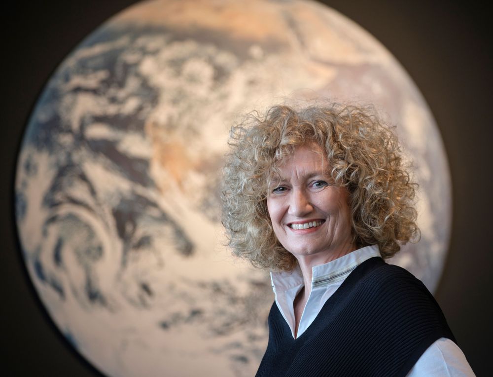 Eine Frau mit lockigem, blondem Haar und einem freundlichen Lächeln steht vor einem großformatigen Bild der Erde. Sie trägt eine weiße Bluse mit einem dunklen Pullover darüber. Der Hintergrund ist unscharf.