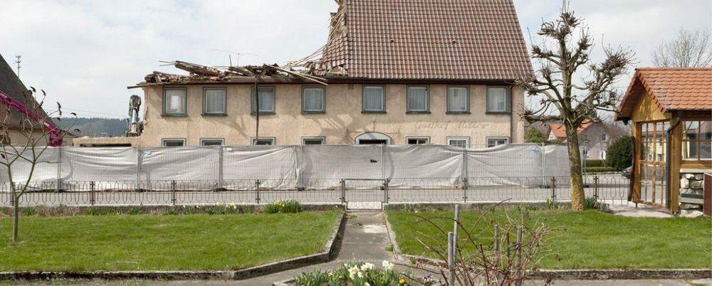 Ein Betonweg führt über einen Rasen zu einem Zaun, dahinter steht hinter weißen Baustellenplanen ein altes Gasthof-Gebäude, bei dem das Dach auf der linken Seiten komplett eingerissen ist.