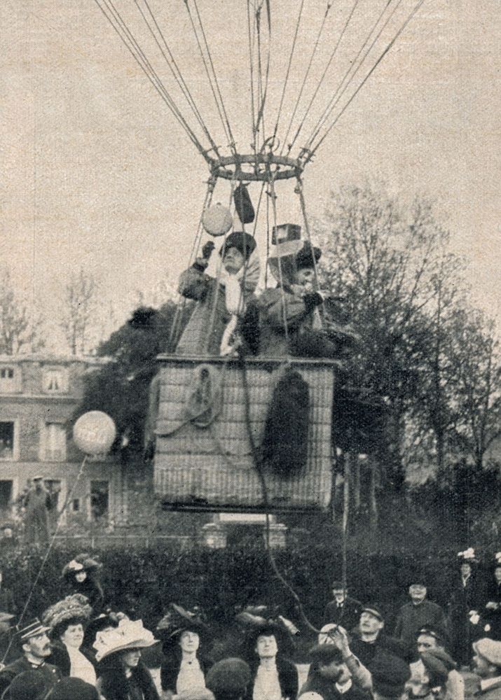 Die historische Aufnahme zeigt zwei Frauen, die im Korb eines aufsteigenden Ballons stehen. Darunter hat sich eine Gruppe von Personen versammelt.