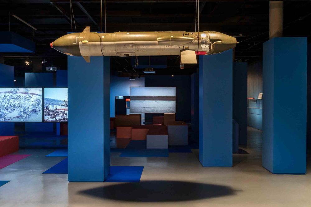 Die Ausstellungsansicht zeigt das Modell eines Lenkflugkörpersystems, welches von der Decke hängt und von de rForm an einen Zeppelin erinnert. Im Hintergrund sind verschiedene Podeste und drei Bildschirme zu erkennen.