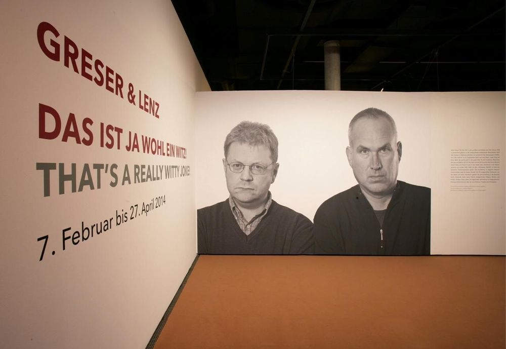 Links an einer weißen Wand befindet sich ein großer Text und auf der mittleren Wand befindet sich ein Porträt von zwei Männern.