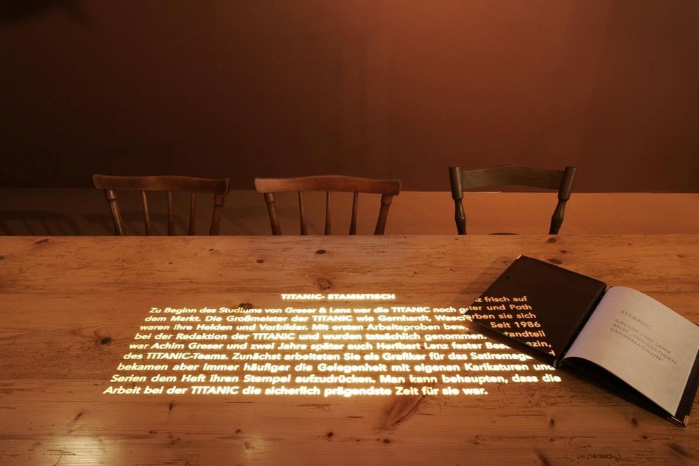 Auf einen Holztisch ist von der Decke ein Text projiziert, neben dem ein aufgeschlagenes Buch liegt.
