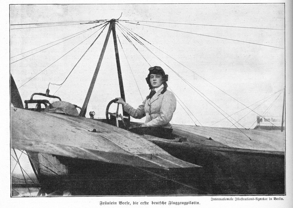 Die historische Aufnahme zeigt eine Frau in Pilotenkleidung, die in einem Flugzeug sitzt und in die Kamera schaut. Sie trägt eine Fliegermütze und hält mit einer Hand das Steuerrad.