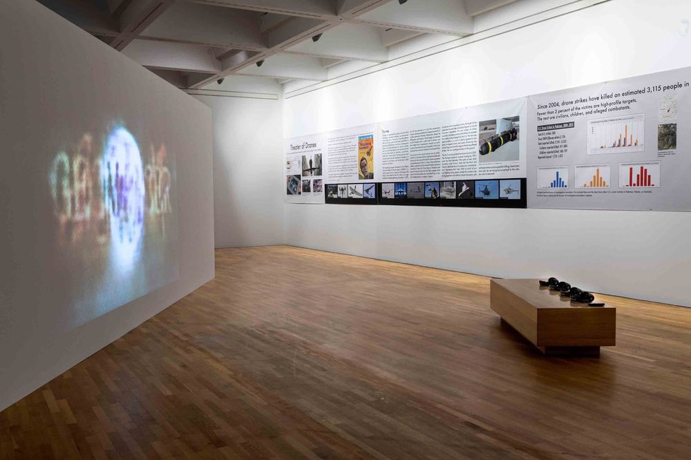 Die Ausstellungsansicht zeigt auf der linken Seiet ein Bild, das an eine weiße Wand projiziert wird. Rechts gegenüber steht eine Sitzbank für Besucher*innen, auf der Kopfhörer liegen. Im Hintergrund an der Wand hängen Bilder und Ausstellungstexte.