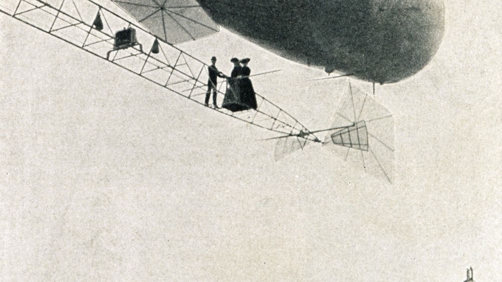 Die historische Aufnahme zeigt einen Mann und zwei Frauen, die in weiter Ferne auf dem äußeren Gerüst eines Luftschiffs stehen. Die drei Personen scheinen dort ohne jegliche Sicherungen zu stehen.