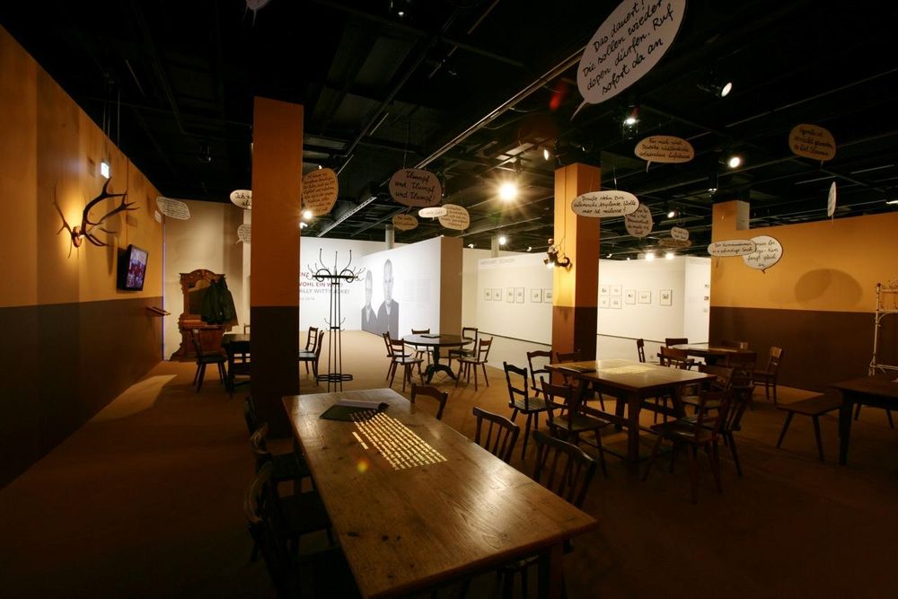 Der Ausstellungsraum ähnelt der Inneneinrichtung eines alten Gasthofes. Um massive Holztische stehen passende Holzstühle und von der Decke baumeln verschiedene Sprechblasen mit Text.
