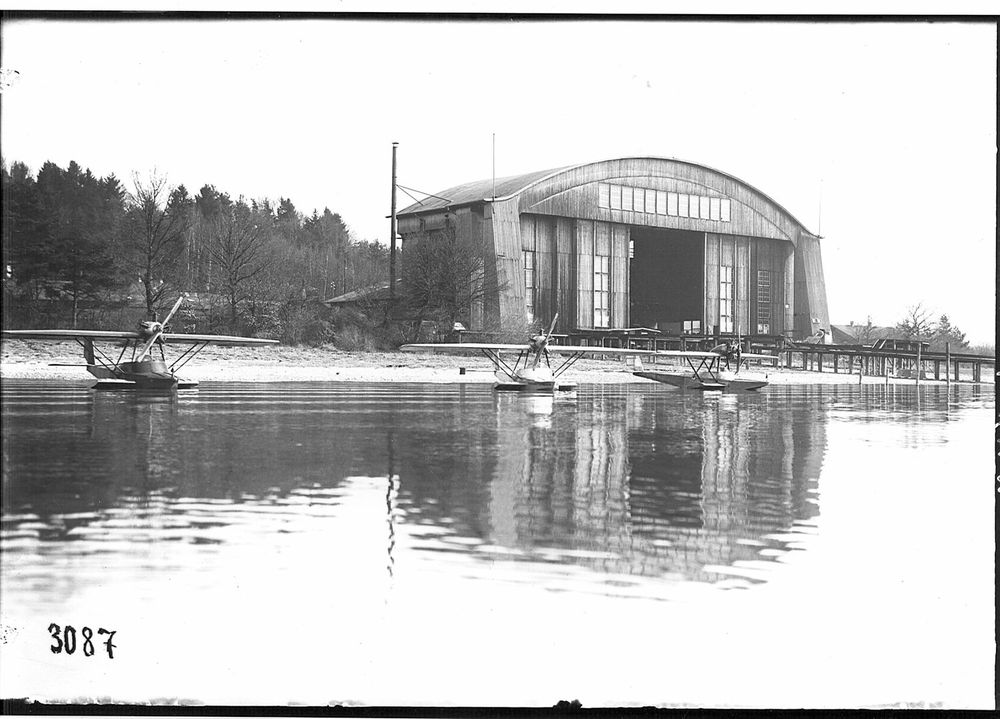 Die historische Aufnahme zeigt drei Dornier Wasser-Flugzeuge vor den großen Werkshallen im Wasser liegend. 