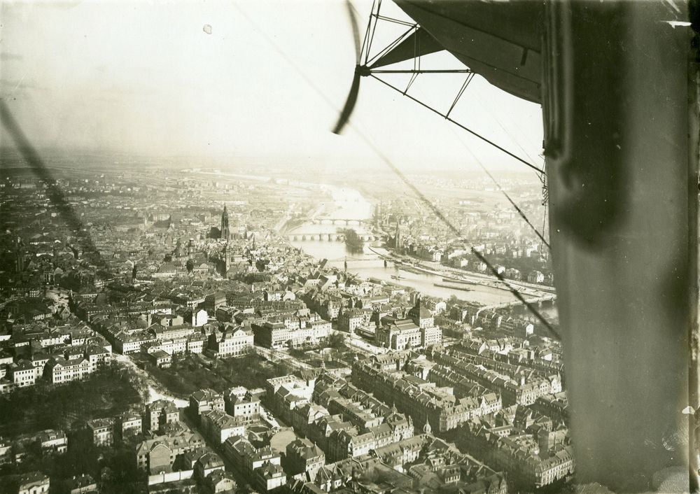 Blick aus einem Delag-Zeppelin auf Häuser in Frankfurt. Im Vordergrund sehr detaillierte Aufnahmen der Häuser. In der Mitte schlängelt sich der Main durch das Bild.
