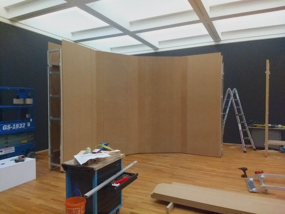 Ein braunes Element einer Ausstellungswand steht mittig in einem Raum mit Holzboden und Kassetten-Decke, die hell beleuchtet ist.