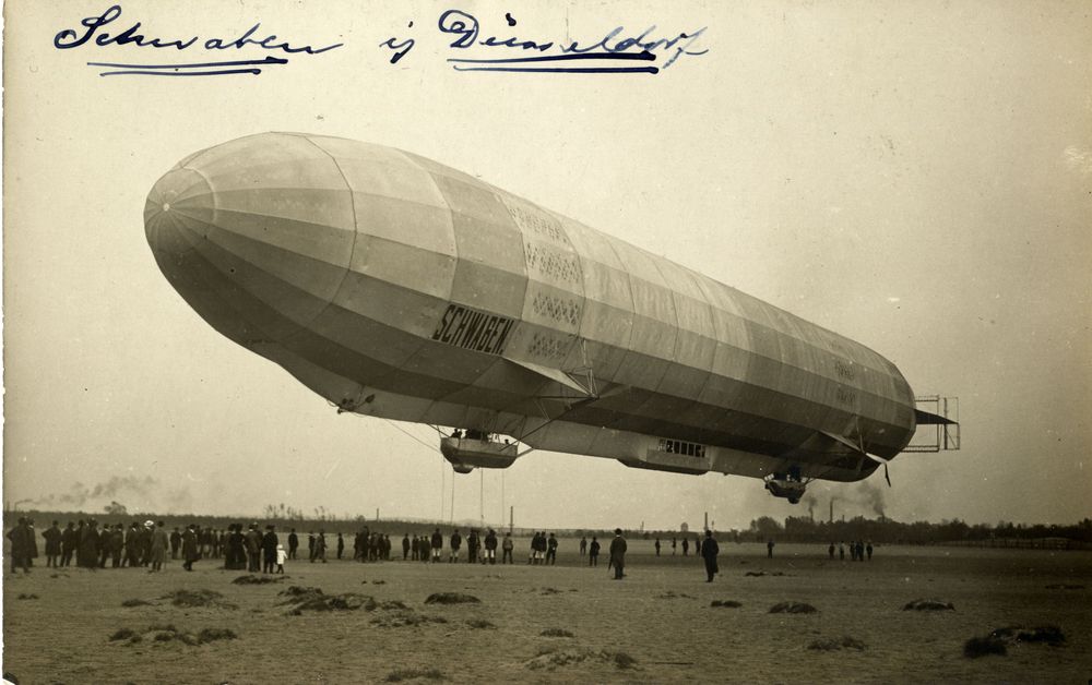 LZ 10 „Schwaben“ schräg von vorne betrachtet, beim Starten über Düsseldorf. Man sieht das Flugfeld mit rund 50 Schaulustigen, aber auch einigen Soldaten, die vermutlich zur Haltemannschaft gehören.