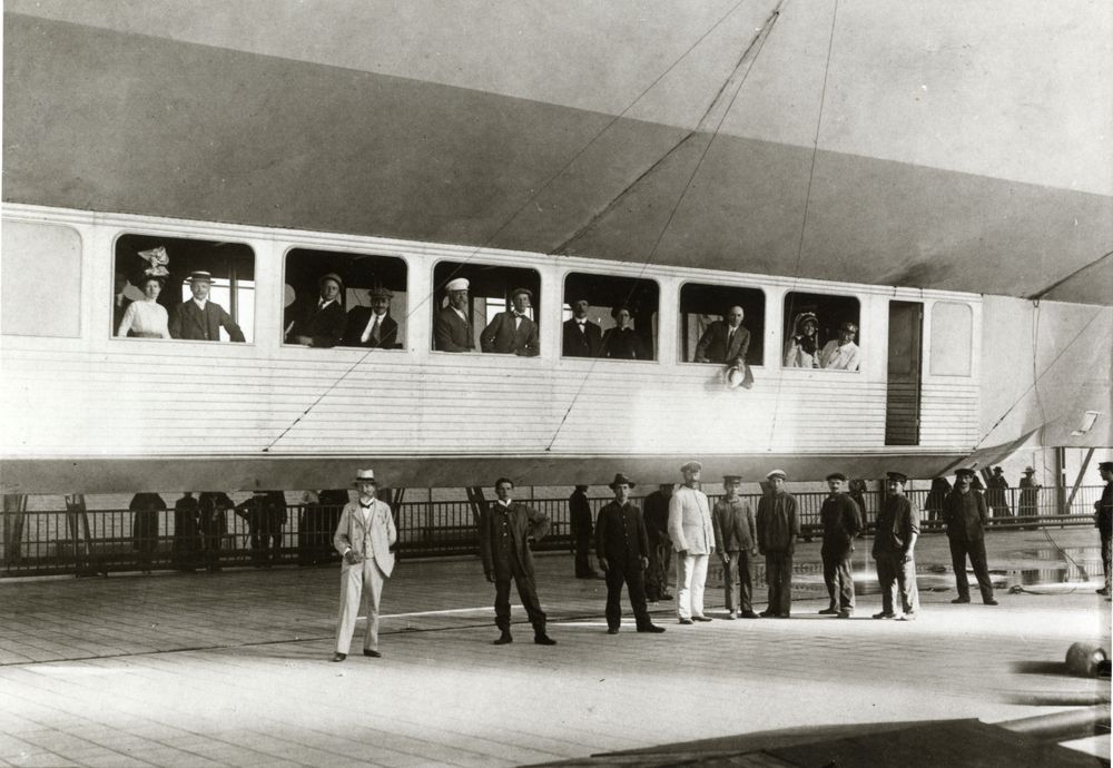 Außenansicht der Passagiergondel der LZ 10 „Schwaben“. Fahrgäste schauen aus den großen Panoramafenstern. Vor der Gondel auf dem Erdboden stehen Arbeiter und Schaulustige.