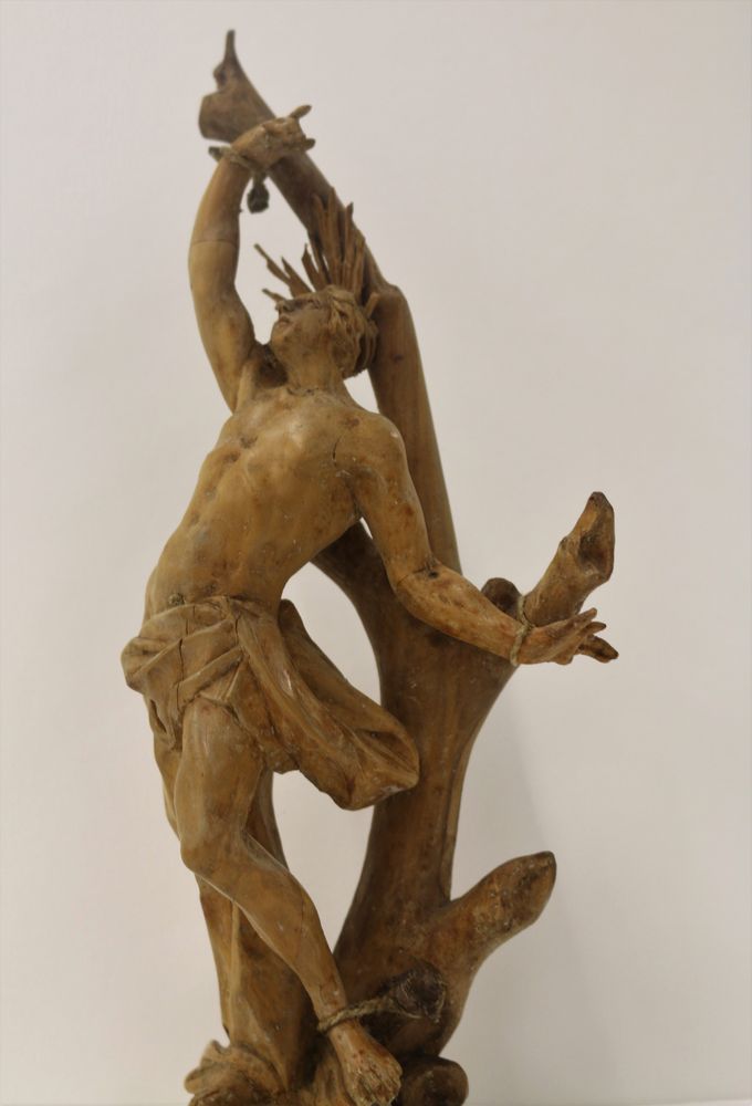 Die Aufnahme zeigt eine Skulptur aus Holz. Die geschnitzte Figur eines Mannes ist an den Hand- und Fußgelenken an den Ast gefesselt. 