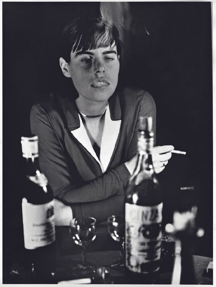 Die schwarz-weiß Fotografie zeigt eine Person mit einer Zigarette in der Hand und Rauch ausatmend. Die Person stützt ihre Elenbogen auf einen Tisch auf dem Gläser und Flaschen stehen.