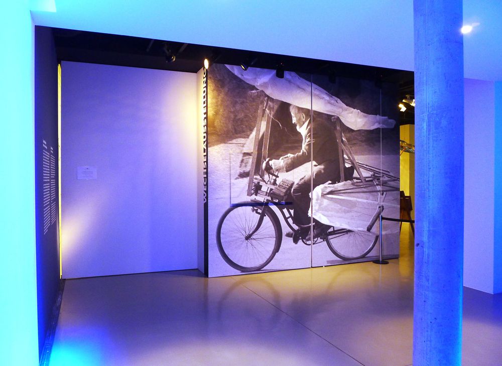 Die Ausstellungsansicht zeigt eine blau beleuchtete Wand, an deren rechten Seite ein großes Bild von Gustav Mesmer hängt, wie er eines seiner Flugräder fährt.