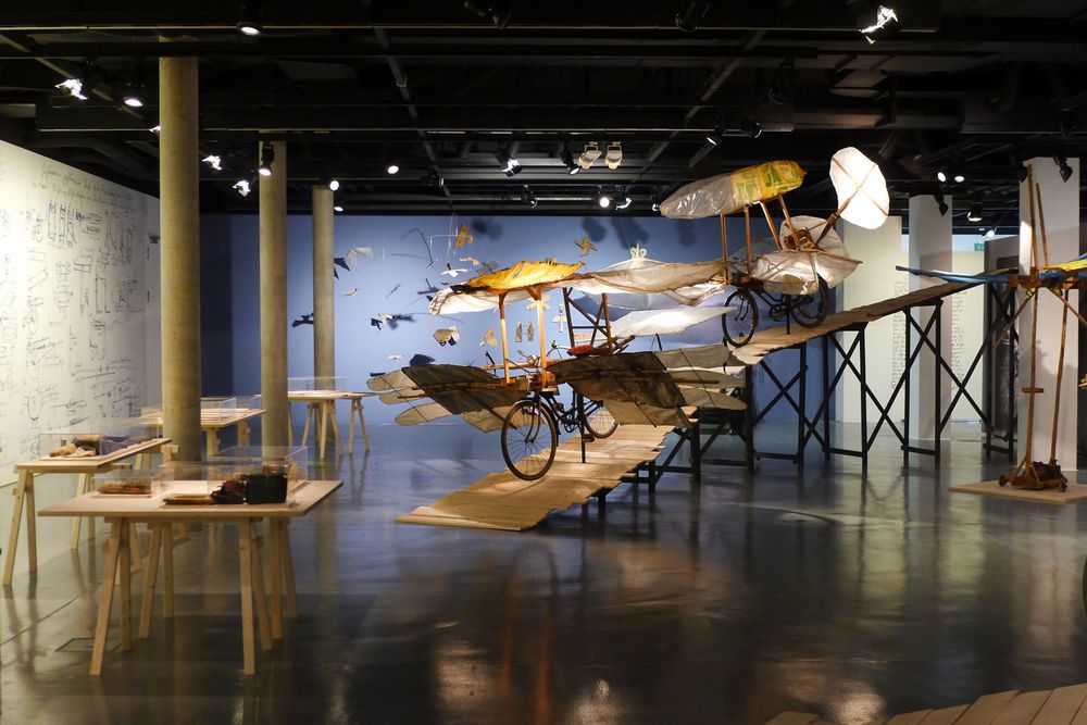 Die Ausstellungsansicht zeigt zwei Modelle von Flugrädern, die auf einer Holztreppe platziert sind. Links im Raum stehen Holztische mit Schaukästen darauf.