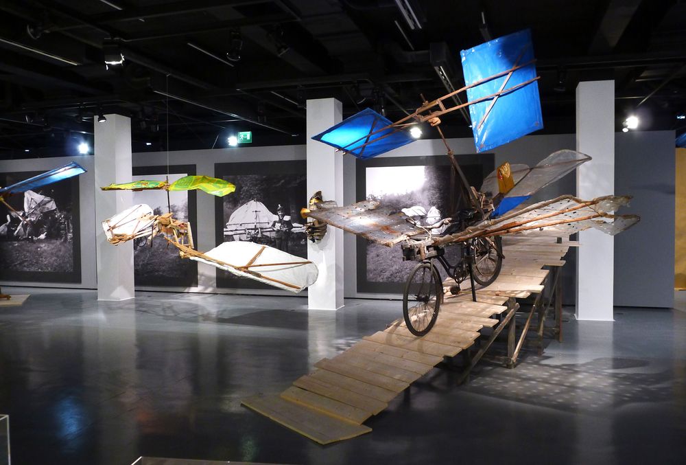 Die Ausstellungsansicht zeigt zwei Modelle von Flugrädern, eines davon steht auf einer Art Holzrampe. Dahinter an den Wänden sind große schwarz-weiß Fotografien angebracht.