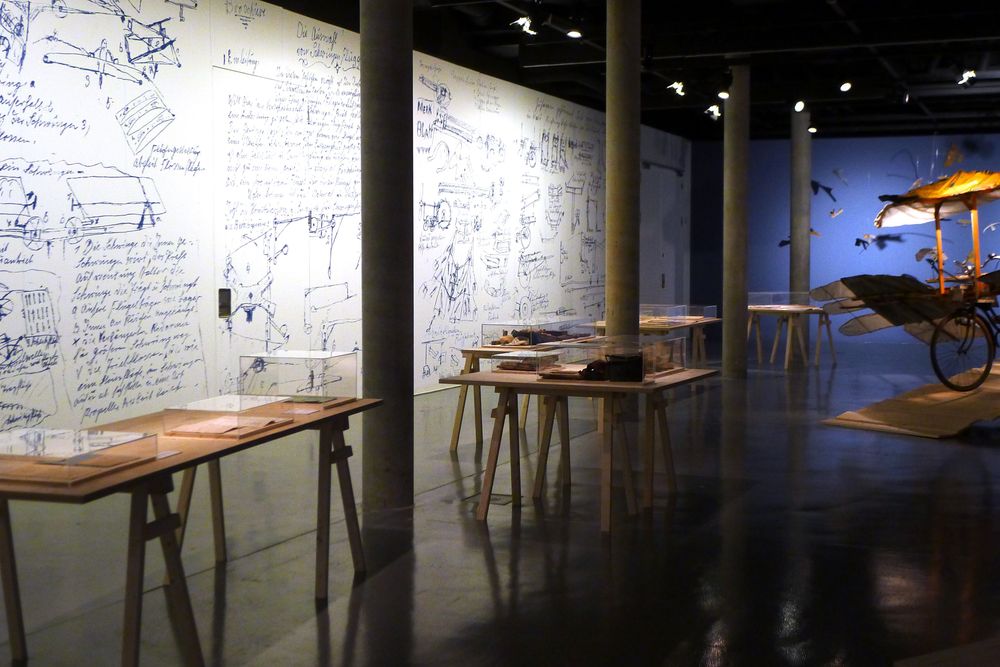 Die Ausstellungsansicht zeigt mehrere Holztische auf denen sich Schaukästen mit Exponaten darin befinden. Die weiße Wand auf der linken Seite ist voll mit großen Skizzen und Texten