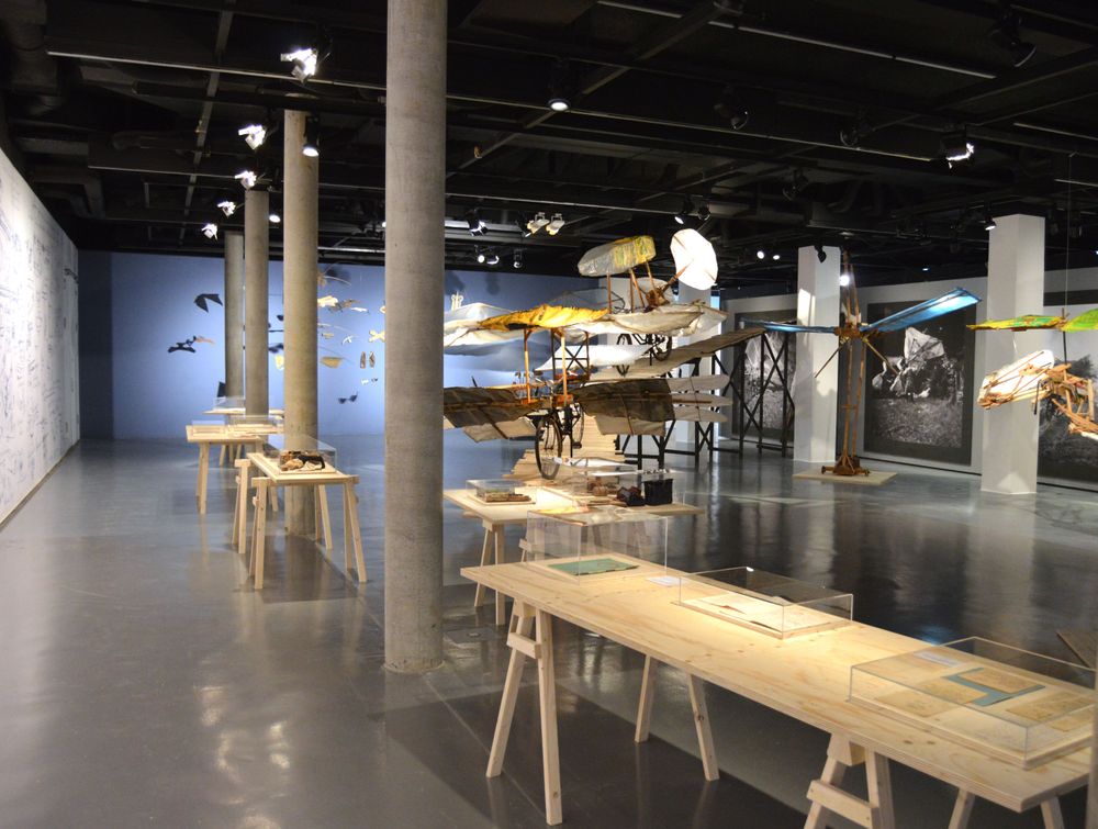 Die Ausstellungsansicht zeigt mehrere Modelle von Flugrädern, die entweder auf einer Holzrampe stehen oder von der Decke hängen. Im Vordergrund des Bildes stehen einige Holztische, auf denen sich Schaukästen mit Exponaten darin befinden.