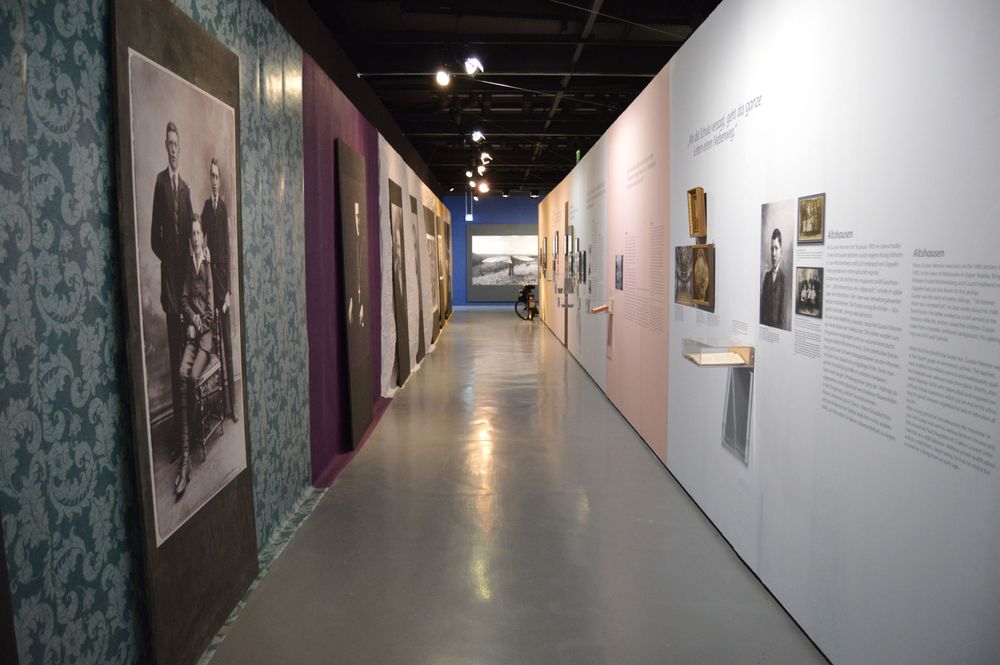 Die Ausstellungsansicht zeigt einen langen Flur, dessen Wände auf der einen Seite mit großen Portraits behangen sind, auf der anderen Seite mit Ausstellungstexten und kleineren Bildern und Objekten.