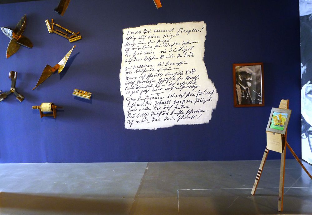An einer blauen Wand befindet sich auf weißem Hintergrund ein handschriftlicher Text, der an einen Notizzettel oder Brief erinnert. Daneben hängen verschiedene Objekte und ein Porträt.
