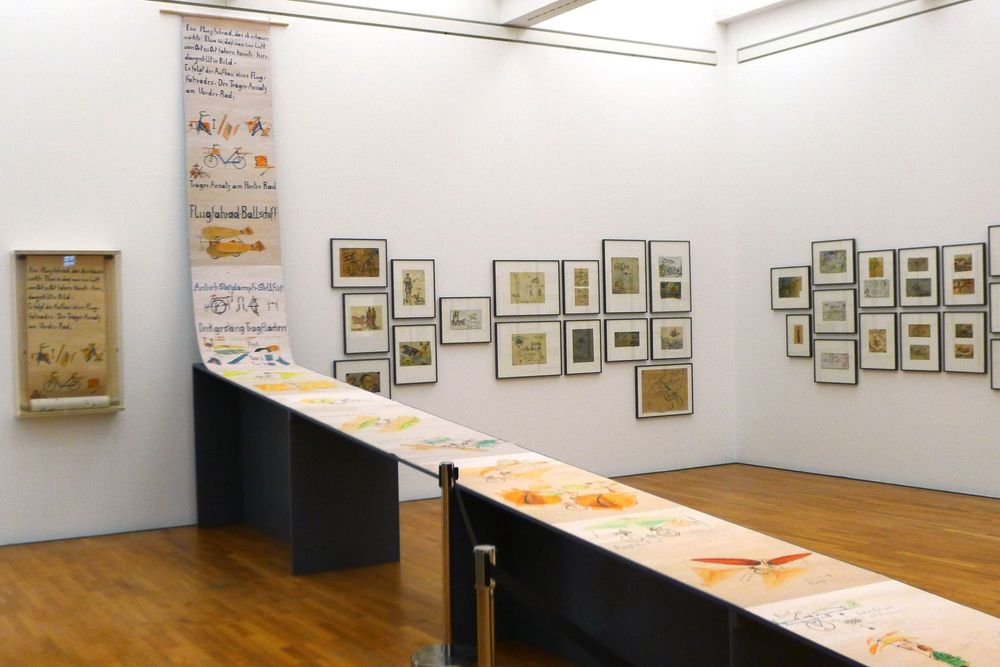 Die Ausstellungsansicht zeigt eine von der Decke über Tische abgerollte Papierrolle, die mit verschiedenen Zeichnungen bemalt ist. Daneben hängen verschiedene Bilder an der Wand.