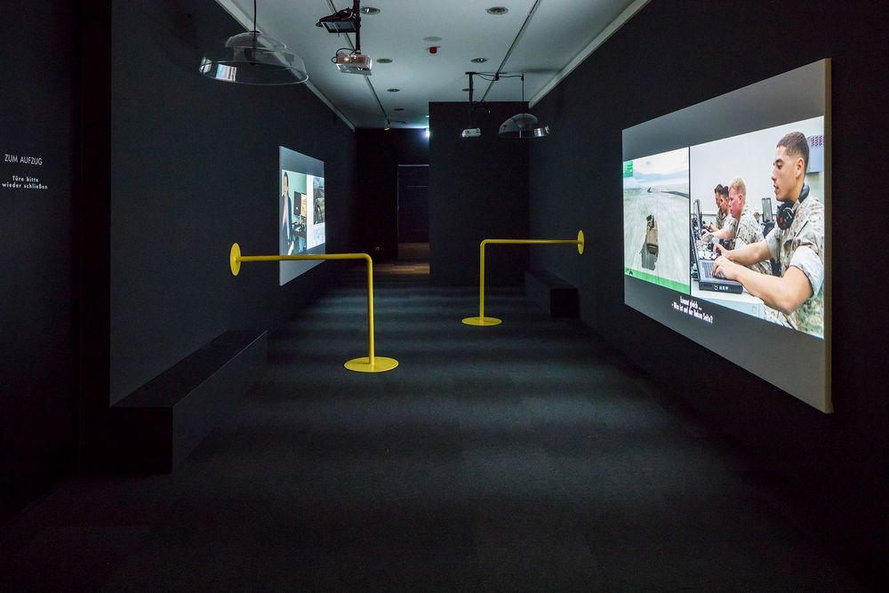 Die Ausstellungsansicht zeigt einen schwarzen, dunklen Flur in dessen Mitte sich zwei grelle, gelbe Geländer befinden. An der rechten und linken Wand projizieren Beamer Videos auf eine Leinwand.