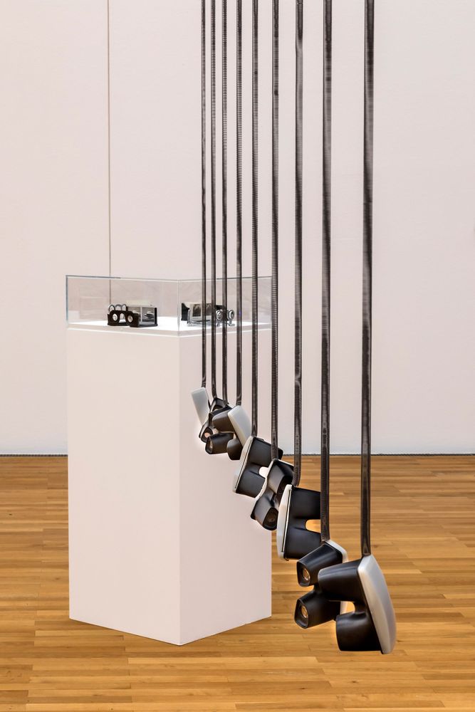 Von der Decke hängen neun identische VR-Brillen, dahinter befindet sich ein weißes Podest auf dem eine Vitrine mit Zeit Ausstellungsobjekten steht. 