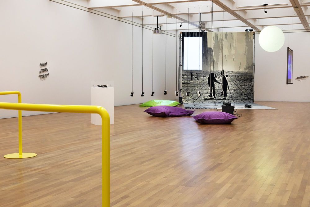 Die Ausstellungsansicht zeigt im Vordergrund zwei gelbe Geländer. Dahinter befindet sich eine Installation die aus mehreren VR-Brillen besteht, die von der Decke hängen. Auf dem Boden bei der Installation liegen drei große Sitzkissen für Besucher*innen.