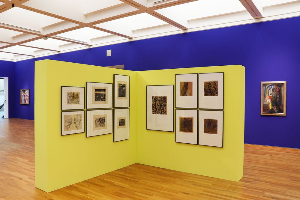 Die Ausstellungsansicht zeigt zwei kleinere gelbe Ausstellungswände mit verschiedenen Bildern daran, die in der Mitte des Raume stehen. Im Hintergrund an einer blauen Wand hängen weitere Gemälde.