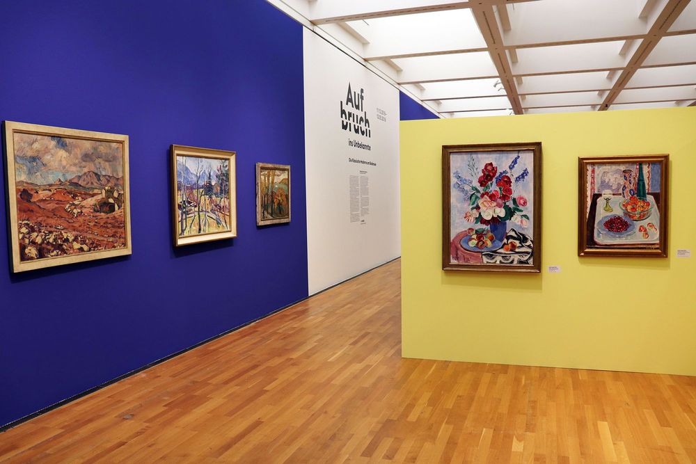Die Ausstellungsansicht zeigt auf der rechten Seite eine gelbe Wand, an der zwei bunte Gemälde hängen. Links an der blau-weißen Wand hängen drei weitere Gemälde und ein Ausstellungstext ist angebracht.